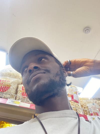 This media file shows a close-up, low-angle, outdoor shot of a person wearing a light-colored cap and a white t-shirt. The person has dark skin and is wearing two silver rings on their right hand, which is reaching up towards their head. They have short, dark hair and a light beard. The background suggests a retail environment, with shelves of bagged popcorn visible. A price tag with the text "N1,820" and "PAROUSIAL POP CORN 550" is also visible. A white smoke detector is mounted on a light-colored ceiling in the upper left of the frame. The lighting is bright, indicating it is likely daytime. The overall mood appears casual and informal.