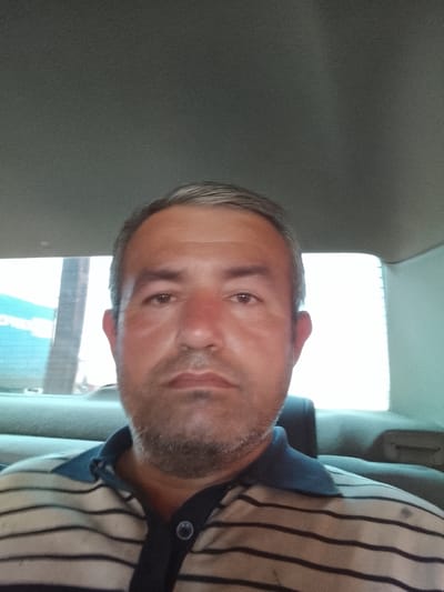 A middle-aged man sits in the backseat of a car. He is wearing a striped polo shirt. The location appears to be Sabir Rakhimov, Uzbekistan based on background details outside the car window. The setting is a car interior with a headrest behind him. The man is looking directly at the camera, suggesting a selfie.