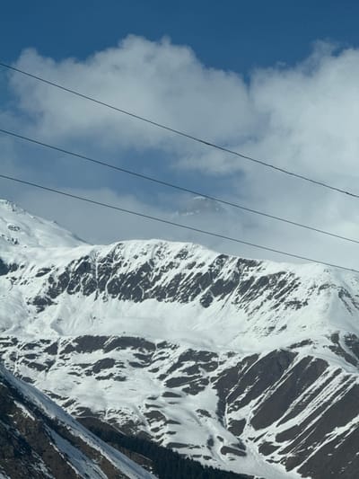 This image depicts a vast, snow-covered mountainous landscape in Kanobi, Georgia. The upper third of the frame shows a clear blue sky with interspersed white cumulus clouds. Four dark, parallel utility lines, consistent with power lines or ski lift cables, traverse diagonally from the upper left to the middle right of the image. The mid-ground and foreground are dominated by rugged mountain peaks and slopes, extensively covered in white snow, with dark rock faces exposed in intricate patterns. Patches of dark green coniferous trees are visible at the base of some slopes in the lower portion of the image. A distant, higher mountain peak is partially visible through a break in the clouds in the upper-mid section of the background. No people or active interactions are visible within the scene. The environment is characterized by a cold, high-altitude, mountainous climate.