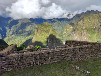 Stone walls frame a landscape of lush, green mountains partly shrouded in clouds. The mountains are a mix of shadowy forests and sunny slopes. A stepped structure of stone is in the foreground, contrasting with the distant peaks. Green grass grows around the base of the stone walls, adding a soft texture to the hard, angular stone. The overall scene conveys a sense of history and natural beauty.