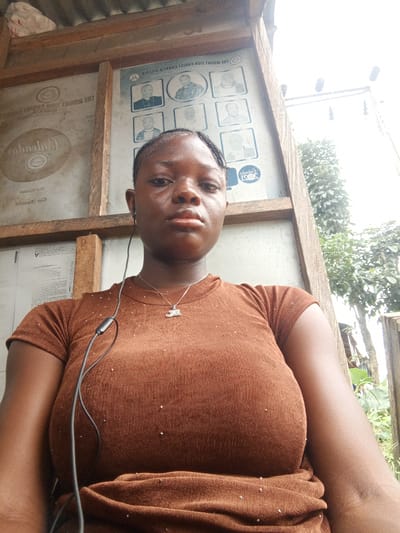 A woman is standing outdoors in Mbribit, Nigeria. She is wearing a brown shirt and a necklace, with earbuds in her ear. Behind her is a wooden structure, possibly a bus stop or shelter, with signage that includes photos of people and text in both English and potentially another language. To the right of the structure, there are trees and glimpses of the outside environment. The area appears to be relatively simple and perhaps rural, though the presence of electrical wires suggests some infrastructure.