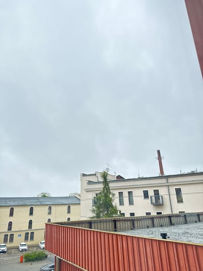 The image captures a view of buildings under an overcast sky, suggesting a cloudy day. The perspective is from a higher vantage point, looking down and to the side at several structures.
On the right side of the frame, a corrugated red roof is prominent, angling downwards. Adjacent to this is a multi-story building with a light-colored facade and a dark roof. This building features numerous windows, some with arched tops, and several doors on its lower levels. A row of parked cars, mostly white, is visible on the pavement in front of this building. Some of these cars are facing upwards, indicating the image is rotated. A small green bush or tree is planted in front of one of the cars. A blue sign with a white 'P' is visible near the parked cars, likely indicating parking.
Further back and to the left, another taller building is visible, also with a light-colored facade and numerous windows. A brick chimney extends upwards from this building, topped with some antenna-like structures. A tree with wispy green foliage grows near this building, partially obscuring some of its windows and balconies.
The overall scene appears to be an urban or industrial setting, possibly a complex of warehouses or apartment blocks, given the architecture and the presence of a parking lot. The lack of direct sunlight and the gray sky indicate overcast weather. The image orientation is noticeably off, requiring rotation for proper viewing. No people are visible in the frame.