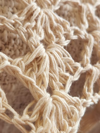The image is a close-up, macro shot of a beige, crocheted textile. The texture is detailed, showing the woven loops and strands of yarn, with some individual fibers slightly frayed. The pattern appears to be a lacy, openwork design. The lighting casts subtle shadows, highlighting the three-dimensional quality of the crochet work. The background is softly blurred, suggesting it is also made of a similar material or a neutral surface. There are no people, animals, or any other objects visible, and the scene is entirely focused on the fabric. The image does not contain any information that can be used to determine its location within Maitland, United States of America, or any other geographical area.