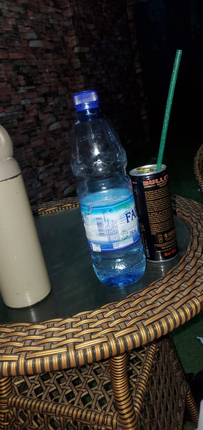 A close-up, eye-level, indoor shot shows a glass table with a bottle of Faro water and a can of Bullet energy drink. The water bottle is plastic and has a blue cap, and the energy drink can is black with red and yellow text. A green straw pokes out of the energy drink can. The background is a brick wall. The image appears to be taken at night. This scene is likely in the city of Numan, Nigeria, given the mention of Faro water, a prominent beverage brand in Nigeria.