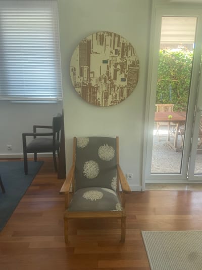 The image shows an indoor scene with a view of an outdoor patio through a large glass door or window. Inside, a wooden armchair with grey cushions decorated with white floral patterns is positioned on a polished wooden floor. To the left of the armchair, a large circular piece of wall art with a repetitive geometric pattern in brown and beige tones hangs on a white wall. Below the wall art, a set of white vertical blinds covers a window. To the right of the armchair, a light-colored rug is partially visible.
The outdoor view reveals a paved patio area with wooden garden furniture, including a table and chairs. Lush green foliage and trees are visible, suggesting a garden setting. A yellow tennis ball rests on the patio furniture. The scene appears to be well-lit, with sunlight streaming in, indicating it's daytime. The overall impression is one of a peaceful and well-decorated living space, possibly a home or a holiday rental in Matosinhos, Portugal.