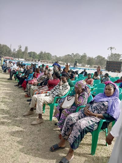 A large group of people sits in green plastic chairs outdoors, likely at an event. Most are seated, facing forward in rows, seemingly listening or watching something. A woman in a patterned dress and purple headscarf is prominently in the foreground, seated with another woman behind her. The ground is grassy and a large structure is visible in the background, possibly a stage. The sky is overcast.