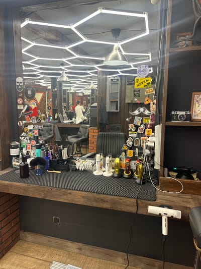 The image depicts the interior of a barber shop, likely located in Lytkarino, Russia, given the context. The scene is dominated by a large mirror reflecting the shop's interior. A barber chair and styling station are visible in the foreground, equipped with various grooming tools such as clippers, brushes, and bottles of hair products. The walls are decorated with numerous stickers, some in Russian, and posters. Overhead, a modern hexagonal lighting system illuminates the space. In the reflection, a barber can be seen attending to a customer. A vintage camera is displayed on a shelf to the right, alongside framed artwork. A white hairdryer is mounted on the wall near the bottom right of the frame. The overall atmosphere suggests a professional and well-equipped barber shop.