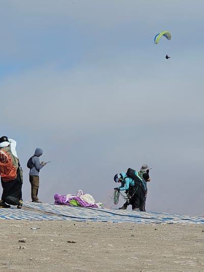 The image depicts a mountainside in Alto Hospicio, Chile, where multiple individuals are engaged in paragliding activities. Near the top of the ridge, three people appear to be preparing for or have just initiated a launch. One is dressed in a blue jacket and dark pants, another in a grey hoodie and tan pants, and the third is wearing dark clothing with orange accents. Two paragliders are airborne in the background. One is a brightly colored wing, while the other is smaller and appears to be a person performing acrobatics without a glider. The sky is a clear blue with some cloud cover. The hillside slopes steeply down to the right.
