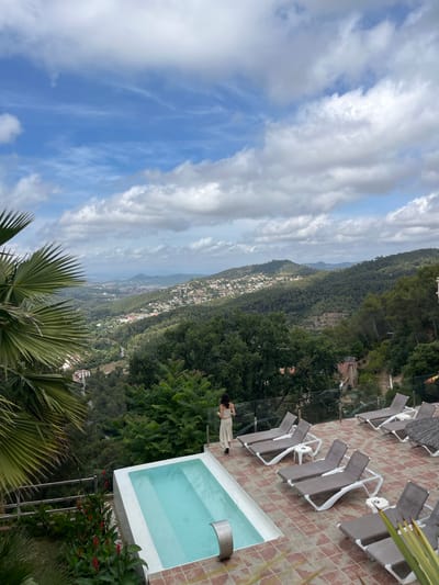 The image depicts a scenic view from a terrace overlooking the town of Can Güell, Spain. A woman, dressed in a light-colored outfit, stands at the edge of a small, rectangular swimming pool, looking out at the landscape. The terrace is paved with terracotta tiles and furnished with several empty lounge chairs. Lush green trees and vegetation surround the terrace, and the distant hills are dotted with houses. The sky is partly cloudy, with a mix of blue sky and white cumulus clouds. The overall impression is one of tranquility and natural beauty.