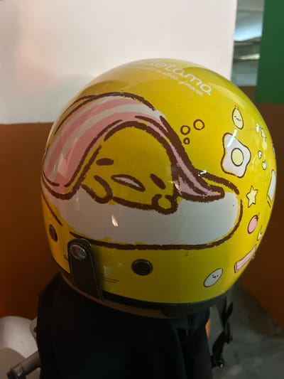 The media file shows a close-up of a yellow helmet decorated with a cartoon character. The character appears to be Gudetama, a lazy egg character from Sanrio, characterized by its unenthusiastic expression and simple design. The helmet has a glossy finish and features various drawings of Gudetama, including its face, and other playful elements like fried eggs and stars. The text "gudetama" and "born to sleep forever" are visible on the helmet.

The helmet is partially obscured by a black object, likely a bag or the wearer's clothing, and is resting on a brown surface, possibly a table or the ground. The background is blurred, but suggests an indoor setting with some hints of green and white. There are metal fasteners and vents visible on the helmet, indicating it is a functional piece of protective gear. The lighting suggests it is daytime, but the indoor setting makes it difficult to determine the exact time of day or weather conditions. The overall impression is of a lighthearted and casual item, possibly used for recreational activities or as a fashion statement. The context of Makassar, Indonesia, adds a regional element to the description, though no specific local landmarks or cues are directly visible in the image.
