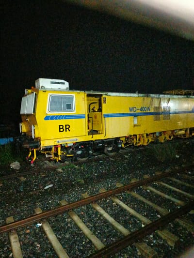 An eye-level, medium shot captures a yellow railway maintenance vehicle, identified by the markings "WD-400W" and "BR," stationary on railway tracks at night in Rajbari, Bangladesh. The vehicle, a prominent yellow color with blue accents and a white upper section, occupies the majority of the frame. Its complex machinery, including various levers, wheels, and a lowered set of tools, is visible beneath its body. The foreground consists of ballast and several sets of parallel railway tracks, with some scattered debris. The background is dark, suggesting nighttime, with a slightly brighter area at the top, possibly from artificial lighting or a distant light source. The scene appears to be an industrial or operational area related to railway maintenance. No individuals are visible in the image.