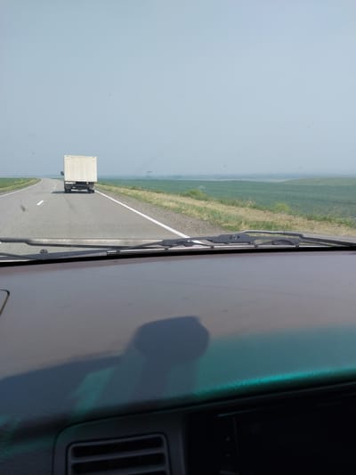 A white box truck is seen from a vehicle's perspective, driving on a paved road. The truck is traveling away from the viewer, in the same direction as the vehicle. The road is divided by a solid white line on the left and a dashed white line on the right. The environment is rural, with green fields on either side of the road stretching to the horizon. The sky is a pale blue and appears hazy. The image is captured through the windshield of the vehicle, with a portion of the dashboard and windshield wipers visible in the foreground. The license plate on the truck appears to read "R449TA".