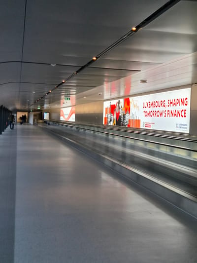 The image shows an indoor moving walkway (escalator) with advertising billboards in what appears to be a tunnel or underground transit area. One billboard prominently displays "LUXEMBOURG, SHAPING TOMORROW'S FINANCE" with an associated cityscape graphic. The area is well-lit with recessed lighting along the wall and is constructed of gray metal panels. People are visible further down the escalator. The location is indicated by the advertising as Luxembourg and likely in Niederanven, Luxembourg due to the image metadata.