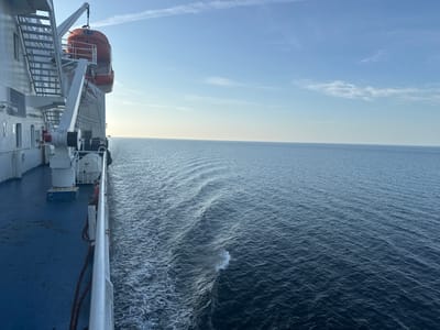 The image depicts the deck of a ship at sea, with the calm ocean stretching out to the horizon under a clear blue sky with wispy clouds. On the left side of the frame, a portion of the ship's superstructure is visible, including metal stairs and lifeboats. The deck is painted blue, and a white railing runs along the edge, separating it from the water. The sea shows gentle waves, creating ripples and a subtle wake behind the vessel. The overall scene is serene and suggests a journey or passage at sea. The location is identified as Unknown, Sweden.