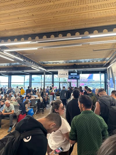 The image depicts an indoor airport waiting area with numerous people queuing near Gate 4. The environment is characterized by a modern ceiling with intricate perforations and overhead lighting. Through large windows, an airplane with a distinctive pink and purple livery is visible on the tarmac, along with jet bridges and other airport infrastructure.

The people in the foreground and middle ground are predominantly dressed in casual attire and are either standing in lines, sitting on benches, or interacting with each other. Several individuals are looking at their phones. Luggage, including backpacks and rolling suitcases, is scattered around the waiting area. A digital display above Gate 4 shows flight information, indicating a destination of Sofia (SOF).

The overall scene conveys a typical airport atmosphere of waiting and departure. The specific location in Künersberg, Germany is not visually evident from the image itself; it is provided as context.