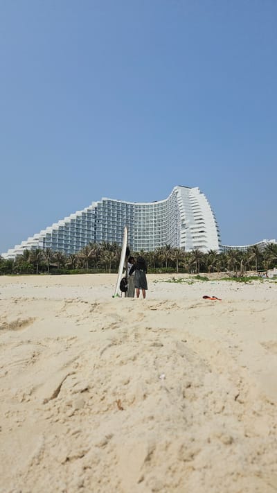 This image captures a vibrant beach scene under a clear, bright blue sky. A prominent, large, white modern building, likely a hotel or resort given its size and design, dominates the background. It features a distinctive curved, wave-like facade with numerous windows and balconies. A dense line of green palm trees runs horizontally, separating the multi-story structure from the sandy beach.

In the mid-ground, two individuals are standing on the light brown sand. One person is dressed in dark attire, while the other, slightly shorter, wears a light-colored top and dark bottoms. Positioned upright between them and slightly in front, is a white surfboard, clearly showing a green fin at its base and a black accessory attached to it. Further along the sandy expanse to the right, a small, reddish-orange object is visible. The scene depicts a sunny day, suggesting good weather for beach activities, consistent with a resort location like Bac Cam Ranh, Vietnam. There is no visible text in the image.