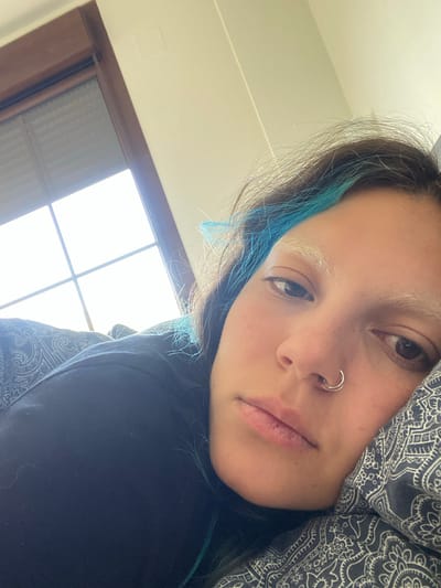 A close-up, indoor shot features a person with fair skin, brown eyes, and a nose ring. The person has dyed blue streaks in their dark hair and bleached eyebrows. They are partially obscured by a gray patterned fabric, likely clothing or a blanket, and wearing a black garment. A window with a grid pattern is visible in the background, allowing bright sunlight to stream in. The setting does not provide sufficient clues to pinpoint a specific location within Villaviciosa, Spain.