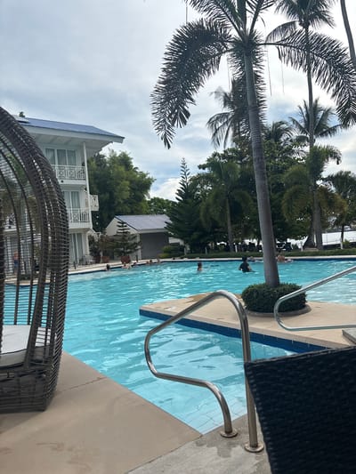 In Samal, Philippines, a resort swimming pool is depicted. The pool is filled with clear turquoise water, and several people are visible swimming and relaxing. A multi-story building with white walls and a blue roof, characteristic of resort architecture, stands to the left of the pool. Palm trees and other lush greenery surround the pool area, suggesting a tropical environment. A woven chair in the foreground partially obscures the view on the left side. Metal railings are installed in the pool for easy access. The sky is overcast.