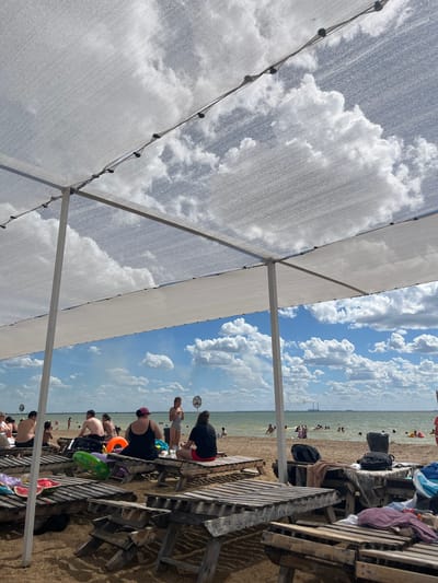 The image depicts a sunny day at a beach with people relaxing and swimming in the water. The foreground shows wooden sun loungers covered with various items, including towels, bags, and a partially cut watermelon. A white canopy supported by poles provides shade over some of the loungers. In the mid-ground, people are sitting on loungers and walking on the sandy beach. Some are in swimsuits, suggesting a recreational activity. The background features a body of water, likely a sea or large lake, with numerous people swimming and wading. Beyond the water, the horizon shows a distant shoreline with industrial structures, identified as belonging to Gres-2, Kazakhstan. The sky is partly cloudy, with fluffy white clouds against a bright blue backdrop, indicating good weather conditions.