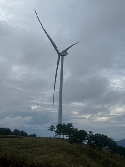 The image shows a wind turbine in Phát Chi, Vietnam. The turbine has three blades and a tall, slender tower. To the right of the turbine are trees and grass. The sky is overcast with grey and white clouds.