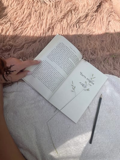 An overhead shot shows an open book lying on a textured pink fluffy blanket and a white terrycloth towel. A person's left hand, with fingernails painted a light pink and a small heart tattoo on the back, rests on the left page of the book, with fingers bent as if holding the page. A dark pencil with small gold star details lies on the right page of the book. The book's pages are white with black text. The right page has a title in Russian that reads "Часть вторая" which translates to "Part two." There is also a minimalist illustration of a plant with seeds. The background is mostly the pink blanket, with a shadow cast across it from the sunlight. The scene appears to be outdoors on a sunny day, likely in a rural setting, given the presence of a thin blade of grass extending from the bottom of the image. The location in the city of Пліскі сельскі Савет, Belarus, is not depicted in the image.