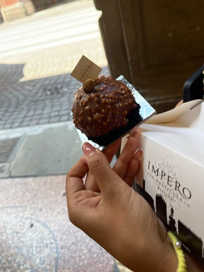 The image shows a person holding a chocolate and nut covered dessert in a square black dish with a brand label in the person’s hand. The dessert appears to be from the Impero Bar Pasticceria in Bologna, Italy, suggested by the branding on the white box. The person is wearing a yellow and white bead bracelet and has pink painted nails. The background shows the city streets of Bologna, Italy. The image is taken in daylight.