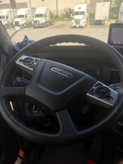 The image shows the interior of a Freightliner truck cab. The steering wheel is the primary focus, with its black textured leather and silver control buttons on either side. The Freightliner logo is visible on the center of the steering wheel. A GPS unit is mounted on the dashboard to the right, displaying a map. Outside the windshield, several white semi-trucks are parked in what appears to be a lot. One of the trucks has a colorful logo on its trailer. The background suggests an industrial or commercial area. The scene is brightly lit, likely during daylight. There are no people visible in the image.