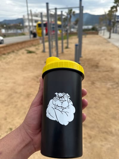 A human hand, with a light skin tone, holds a black cylindrical shaker bottle with a bright yellow ribbed lid in the foreground. The bottle features a white graphic of a bulldog's head. The scene is an outdoor calisthenics park located in Port D'andratx, Spain.
The background, blurred, depicts an outdoor public exercise area with multiple silver metal pull-up bars and exercise structures embedded in a sandy or dirt ground. Behind the exercise equipment, a paved road or promenade runs horizontally. Vehicles are partially visible on the road, including a yellow vehicle resembling a bus. Beyond the road, a body of water, consistent with the Mediterranean Sea, stretches to the horizon, where distant mountains or hills rise under an overcast sky. Several palm trees are sparsely distributed throughout the blurred background.