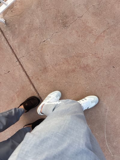 A high-angle shot depicts two pairs of feet standing on a textured, reddish-brown concrete surface. The left foot is shod in a black casual shoe, while the right foot wears a white sneaker with grey stripes. Both feet are attached to legs clad in grey trousers. The lower left corner of the frame shows a fragment of white metal railing. The location is specified as Tabaroucht, Morocco, although there is no direct visual evidence in the image to confirm this. The scene is still, with no apparent action or interaction.