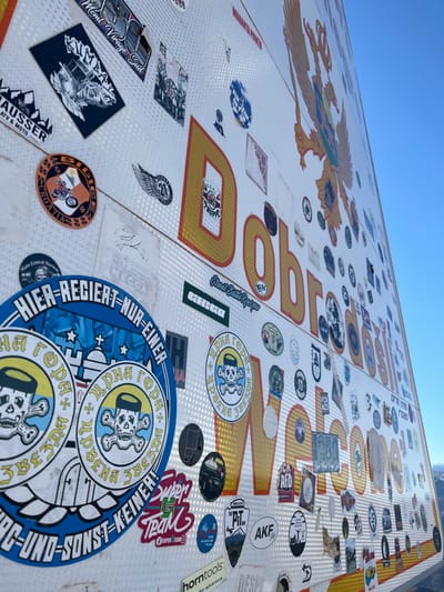 The image displays a large, reflective white surface, likely a roadside sign or monument, situated outdoors under a clear blue sky. The surface is extensively covered with numerous stickers of varying sizes, shapes, and designs. Prominently featured in large, golden-yellow, outlined letters are the words "Добродошли" (Dobrodošli, meaning "Welcome" in Cyrillic) and partially visible "Welcome" in Latin script below. A large golden double-headed eagle with a shield on its chest, consistent with national symbols of the region, is affixed to the upper right portion of the sign.
The stickers present a diverse collection, primarily associated with motorcycle clubs, rallies, and automotive interests. Notable examples include "BIG RALLY CROATIA 06," "MOTO VINTAGE SUD," "CHAUSSER ATV & MOTO," "BOSS TRUPPE," "Street Gang Rodrigo," and "Super G Team." Two prominent circular blue stickers in the lower-left feature a skull wearing a traditional cap, crossed bones, and Cyrillic text "ЦРНА ГОРА" (Crna Gora, Montenegro), along with German text "HIER-REGIERT-NUR-EINER" ("Only one rules here"). Many smaller, less legible stickers also contribute to the dense coverage. The background features a clear blue sky in the upper right quadrant, with a blurred, distant landscape or mountains visible along the far right horizon. This scene is located in Klobuk, Bosnia and Herzegovina.