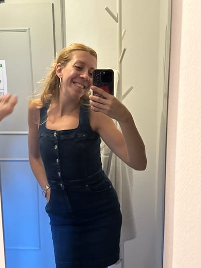 A woman is taking a selfie in a mirror. She has blonde hair and is wearing a dark denim dress with buttons down the front. She is smiling and has a hand raised to the camera. The background appears to be a dressing room or bathroom with a white towel hanging on a rack and a textured white wall. There is a blue-tinted door or cabinet to the left of the frame. The image has a slight yellowish cast. The location is not specified in the image or context.