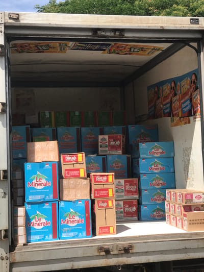 The inside of a truck is filled with boxes of drinks. The brand "Le Minerale" is prominent on blue boxes stacked throughout the truck. There are also brown boxes labeled "Teh Pucuk Harum." The inside walls of the truck have posters advertising various food and drink products, including "Frutta Gummy" and "Durian Gelato." The overall environment appears to be a delivery or storage space for beverages, with the goods packed tightly inside the truck. Given the brands and the OCR, it is likely that the truck is in the city of Miruektaman, Indonesia.