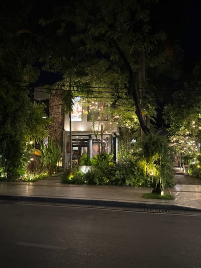 The image captures a nighttime street view of a restaurant facade in Siem Reap, Cambodia. The building is adorned with numerous string lights that illuminate the lush greenery surrounding it. The facade features a stone-clad pillar and a large poster on the upper level. A sidewalk runs in front of the building, bordered by a street. The atmosphere is warm and inviting, with the lights casting a soft glow on the plants and architecture.