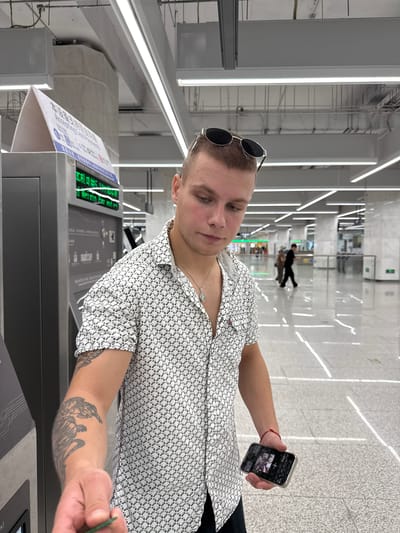 In the Liwan District of China, a young man with a tattoo on his left arm and sunglasses on his head is standing in front of a ticket machine. He is wearing a white short-sleeved shirt with a black geometric pattern and a silver necklace. He is holding a green card in his left hand, reaching towards the ticket machine. In his right hand, he is holding a smartphone displaying a picture. The environment appears to be a modern, well-lit subway station with polished floors and fluorescent lighting. Other people are visible in the background, walking through the station.