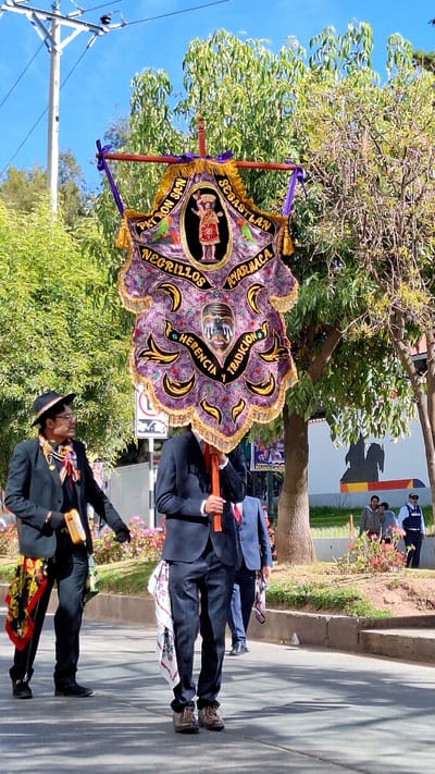 Two men in dark suits are walking in a procession on a paved street. The man on the left wears a black hat and glasses and carries a yellow object. Both men have brightly colored scarves tucked into their suit jackets, one red and yellow patterned, the other white with a red and black pattern. They are carrying a large, elaborately decorated banner with a fringed border. The banner is purple with gold embroidery and depicts various figures and text, including what appears to be a person in a headdress and a mask. Trees and a utility pole are visible in the background, along with a white wall with a mural. Further in the background, several people are standing and observing the procession. The event appears to be taking place in Wanchaq, Peru.