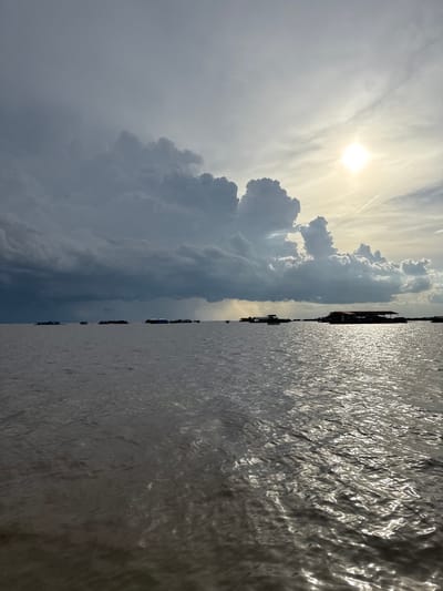 The image depicts a wide expanse of water, likely a lake, with a village of floating houses visible in the distance. The sky is dramatic, with dark, heavy clouds dominating the left side of the frame, suggesting an impending storm or recent rainfall. The sun is visible on the right, breaking through the clouds and casting a bright glare on the water's surface, creating shimmering reflections. The water itself appears calm, with gentle ripples. The floating village consists of several structures, some appearing to be dwellings or businesses, built on rafts or platforms. The overall atmosphere is one of natural beauty and the resilience of a community living in harmony with its aquatic environment. This scene is consistent with the floating villages found on Tonle Sap Lake, a significant geographical feature near Siem Reap, Cambodia.