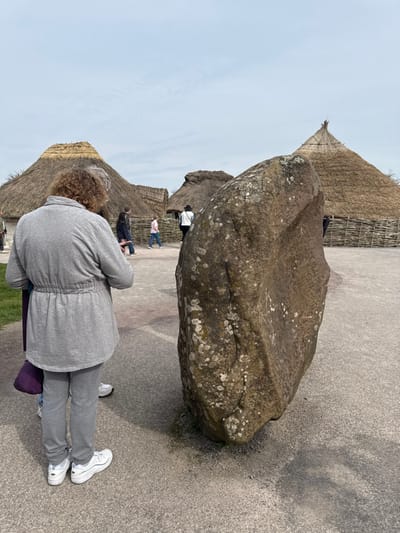 The image captures an outdoor scene at the Neolithic Village reconstruction, located near the Stonehenge Visitor Centre in Amesbury, United Kingdom. In the foreground on the right, a large, irregularly shaped standing stone, dark grey and covered in patches of white and green lichen, stands vertically on a light grey, compacted ground surface. To the left of the stone, a person with curly brown hair, seen from behind, wears a light grey zipped jacket with a cinched waist, matching grey trousers, and white athletic shoes. A purple bag is partially visible by their left leg.
In the midground, several reconstructed roundhouses with conical thatched roofs and wattle-and-daub walls are visible under an overcast, light grey sky. A low woven fence separates a pathway from some of these structures. Multiple other individuals are present in the midground, walking or standing. One person in a white top and dark hat stands near a roundhouse, while another in a pink top and blue trousers walks from left to right along a pathway near the fence. The overall environment is an open-air historical site with scattered trees visible in the distance.