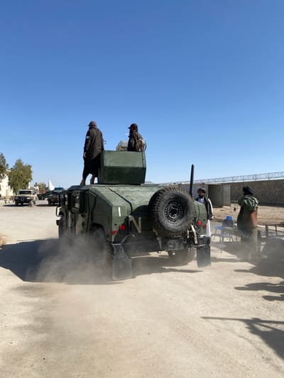 This image depicts an outdoor scene in Mandisar, Afghanistan, under a clear blue sky. A dark green military-style utility vehicle, resembling a Humvee, is positioned in the mid-ground, angled with its rear towards the viewer, moving right. Dust is visibly emanating from its rear wheels, indicating motion on a dry, unpaved surface. The vehicle features a rear-mounted spare tire, "N" painted in white on its left rear, and a vertical white stripe. Two individuals are on top of the vehicle: one stands on the left side of a roof-mounted structure, wearing a dark, long garment and head covering with a light emblem on the left arm; the second individual is seated or crouched within the roof structure, wearing a dark jacket, cap, and a backpack, facing right. To the right of the moving vehicle, two additional individuals are standing on the ground. One wears a white tunic, dark vest, and dark turban, facing left. The other, further right, wears dark clothing with a light wrap. Two blue plastic chairs are visible behind them. In the left background, a paved road leads to several light-colored pickup trucks and other vehicles. Bare trees and light-colored buildings are also present on the left. The right background features a concrete wall topped with razor wire, behind which temporary structures or containers are visible. Strong shadows indicate bright sunlight.