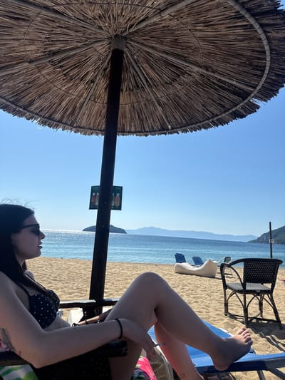 A young woman sits in a lounge chair on a sandy beach, facing the sea. She is wearing a black polka dot bikini and sunglasses. Her arms are resting on the chair, and her legs are extended. A cigarette is held between her fingers. Behind her, the sea is calm and blue, with a small island in the distance. A straw umbrella shades her from the sun. Other beach furniture, including chairs and a beanbag chair, are visible on the sand. The sky is clear and blue. The location is Nea Iraklitsa, Greece.