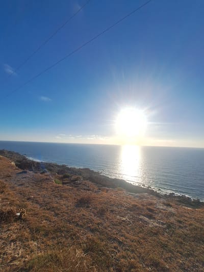 This media file depicts a sun-drenched coastal scene. The primary subjects are the bright, radiant sun, the vast expanse of the ocean, and a rugged, dry, grassy cliffside. The setting is clearly a natural landscape, likely a viewpoint overlooking the sea.

The sun is positioned high in the sky, casting a powerful glare and creating a shimmering reflection on the water's surface, indicating it is likely midday or late afternoon when the sun is at its most intense or beginning its descent. The sky is a clear, vibrant blue, with only a few wisps of white clouds visible in the distance.

The cliffside is covered in dry, golden-brown grasses and sparse vegetation, suggesting a warm or arid climate. The rocky texture of the cliff face is also apparent, adding to the ruggedness of the terrain. The ocean is a deep blue, with gentle waves lapping at the base of the cliffs. The overall impression is one of natural beauty and serenity, with the intense sunlight contributing to a feeling of warmth and openness.

There are no people or specific objects readily identifiable, other than what appears to be power lines running diagonally across the blue sky in the foreground of the image. The composition and lighting suggest a focus on the natural elements and the grandeur of the coastal landscape. No visible text is present in the image. The location context provided is Ahrara, Morocco, which aligns with a coastal, sunny environment.