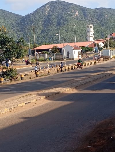 This photograph captures a dynamic street scene in Namanga, Kenya, set against a magnificent backdrop of a large, verdant mountain. The image appears to be taken during either early morning or late afternoon, as suggested by the long, pronounced shadows cast across the road and the warm, golden sunlight illuminating the scene. The sky is clear and blue, indicating fair weather.
The primary subjects include several people engaged in daily activities. Individuals are seen walking along the central median and sidewalks, while others are seated on the yellow-and-black painted bollards lining the road. Two motorcycles are visible; one is being ridden on the road, while another is parked or being prepared for departure.
The scene features a wide paved road, flanked by concrete sidewalks and a central divider marked by numerous yellow bollards. On the right side of the road, a cluster of buildings stands behind a fence. Most notably, a distinctive white building with a multi-tiered tower, reminiscent of a church or public institution, dominates the skyline on that side. Adjacent to it are other structures with reddish-brown roofs and a smaller white guardhouse near the entrance. Several lampposts and utility poles are distributed throughout the scene. Lush green trees and vegetation are scattered along the road and adorn the mountain.
The towering mountain in the background is densely covered with trees, showcasing its rugged topography. The overall impression is one of everyday life unfolding in a semi-urban environment, harmoniously integrated with the natural landscape. No legible text is discernible on any signs or objects within the image.