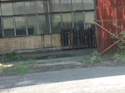 This image captures an industrial scene in Zhivkovo, Bulgaria, characterized by a sense of age and partial disuse. The foreground features an asphalt or gravel surface, partially shadowed, leading to a patch of overgrown grass and weeds.
In the mid-ground, a weathered concrete foundation supports a large industrial building, likely a factory or warehouse. The building's upper section is dominated by multiple large, rectangular windows, possibly frosted or dirty, reflecting some of the hazy daylight. Dark stains run vertically down the concrete foundation beneath the windows.
To the right of the building, a collection of dark-colored gas cylinders is stored outdoors in several metal racks or cages. These cylinders appear old and weathered. Adjacent to these cylinders and extending into the right side of the frame is a heavily rusted metal fence, constructed from chain-link mesh and square-tubing frame. Small, leafy plants and weeds grow both along the base of the building and through the rusted fence.
The scene suggests a daytime setting with visible sunlight and shadows. There are no people visible, and no discernible activity is taking place, contributing to an atmosphere of static industry or quiet neglect. No text is visible in the image.