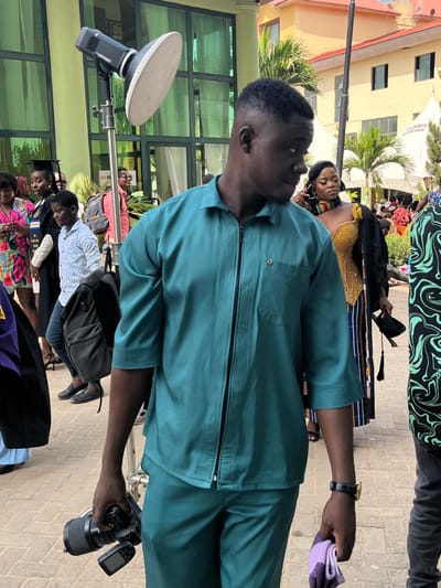 This image depicts an outdoor gathering in Madina, Ghana, featuring a primary subject and several individuals in the background.
The central figure is a dark-skinned adult male, positioned slightly right of the frame's center, facing towards the viewer's right. He wears a short-sleeved, collared, teal-colored shirt with a black zipper closure down the front and matching teal trousers. His right hand holds a black DSLR camera with a lens attached, while his left hand holds a folded purple and white cloth. A black watch with a gold-colored bezel is visible on his left wrist.
In the immediate background to the left of the central figure, a silver-colored studio strobe light with a white circular reflector is mounted on a black stand. Behind the strobe light and further left, a modern building with light green columns and large green-tinted glass windows is visible. A dark-skinned adult female in a black graduation gown and cap, a young dark-skinned boy in a light-colored plaid shirt carrying a black backpack, and another woman in a colorful patterned dress are visible in this area.
To the right of the central figure, a dark-skinned adult female is visible, wearing a gold-colored, possibly beaded, strapless top with a black and striped skirt, and a multi-colored beaded choker necklace. She holds a black graduation gown and cap. Further right, a person in a black and green patterned shirt is partially visible.
The distant background includes a two-story building with an orange-yellow facade and a brown roof, several green palm trees, and multiple white event tents, one bearing "TV EVENTS" branding. Numerous other individuals, some in academic regalia, are present throughout the background. The ground is paved with light brown interlocking bricks. The overall scene suggests a formal outdoor event, possibly a graduation ceremony.