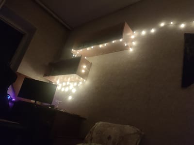 The image shows a dimly lit room, likely a bedroom or living space, with a focus on decorative string lights. The lights, which are small and round, are draped along the top edge of two floating shelves that are mounted on a textured, light brown wall. One shelf is directly above the other, and both are angled upwards from left to right. The shelf on top appears to be wider and deeper than the one below it. The room is mostly dark, with the warm glow of the string lights providing the primary illumination.

In the foreground, to the left, a black computer monitor is visible on a dark desk or dresser. To the left of the monitor, a person's head is partially visible, wearing headphones with blue and purple lighting accents. The person is silhouetted against the ambient light. Below the monitor and desk, a dark, indistinct area suggests more furniture or clutter. In the lower right corner, a section of a pillow or cushion with a patterned fabric is visible.

The overall mood is cozy and ambient, created by the soft lighting and the informal arrangement of the shelves and lights. The presence of the computer equipment suggests a living or working space. The scene appears to be indoors, and the time of day is likely evening or night due to the darkness and the use of decorative lighting. There is no visible text in the image. The location context is Kamianka-Dniprovska, Ukraine.