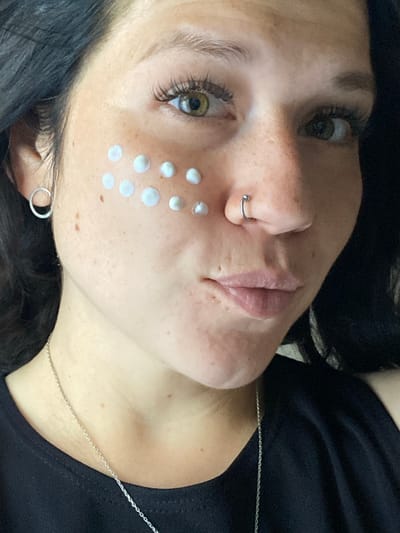 The image shows a close-up, slightly angled view of a woman's face, from the nose up, with a portion of her neck visible. She has dark hair, green eyes, and light skin with freckles and some areas of vitiligo. She is wearing a silver hoop earring in her left ear and a silver nose ring. Several small dots of a white cosmetic product, likely sunscreen or moisturizer, are applied to her cheeks. She is wearing a black top and a silver necklace. The background is dark and out of focus, suggesting an indoor setting. There is no discernible indication of the location being Chaikovsky, Russia.