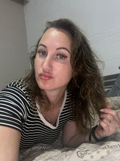 A woman with brown hair and blue eyes lies on a bed. She is wearing a black and white striped shirt and has makeup on. Her lips are parted. She is holding her hair in her right hand. The background appears to be a wall and a bed. There is no indication of the city of Zarichne, Ukraine.