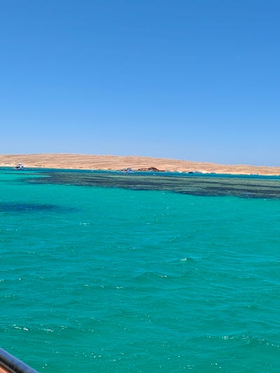 This is a vertical, eye-level, wide shot of the Red Sea near Hurghada, Egypt. The image is divided into three horizontal sections: the sky, the sea, and the land.

The top third of the image is a clear, bright blue sky with no clouds.

The middle section features the sea, which is a vibrant turquoise color, transitioning to a darker blue in some areas due to depth or shadows from the reef. A visible coral reef runs horizontally across the frame, appearing as a dark, irregular line beneath the surface of the water. Several boats are scattered across the water, some white and some blue, appearing to be stationary or moving slowly.

The bottom third of the image shows a sandy, arid coastline. The land is characterized by rolling hills of light brown sand, with sparse vegetation or rock formations visible. Some of these formations are brown and rocky, contrasting with the sand. A metal railing, likely part of a boat, is visible in the lower left corner of the frame, suggesting the photo was taken from a vessel.