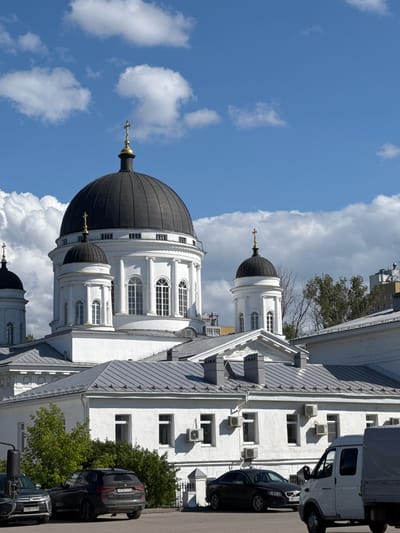The image shows the Ascension Cathedral, also known as the Alexander Nevsky Cathedral, located in Nizhny Novgorod, Russia. The cathedral features a large, black dome topped with a golden cross, and smaller domes on surrounding structures, also capped with crosses. The cathedral is painted white and has a classical architectural style with arched windows. In front of the cathedral, there is a white building with a grey roof. Several cars are parked on a street in the foreground, including a black SUV, a black sedan, and a white van. The sky is blue with scattered white clouds. The scene depicts a sunny day in a city with a prominent religious landmark.