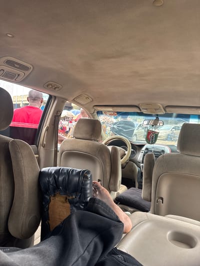 The image shows the interior of a car, likely a minivan given the multiple rows of seats. The view is from the passenger seat, looking towards the front of the vehicle and out the windshield. Two people are visible. One is the photographer, whose legs and arm are in the foreground, wearing dark pants and a black glove. The glove appears worn and ripped, exposing wood underneath. Another person, wearing a red shirt, is partially visible in the back seat. Through the windshield, a street scene is visible with people and market stalls, suggesting an outdoor environment. The lighting suggests it is daytime. The car's interior is upholstered in a light-colored fabric, with visible air vents and a rearview mirror. The windshield has several cracks. The overall scene appears to be in Orji, Nigeria, based on the provided location context, and the image captures a candid moment within a vehicle, possibly during a stop at a marketplace.