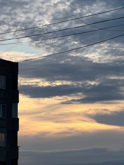 A tall, dark, concrete building stands to the left of the frame, its facade marked by rows of windows and projecting balconies. The sky above is filled with clouds, ranging from light grey and white to darker, stormier hues. A band of warm, golden light illuminates the horizon, suggesting either sunrise or sunset. Several electrical wires are strung across the upper portion of the image, cutting through the cloudy sky. A small bird is visible in flight against the bright horizon. The scene appears to be an urban environment, likely in Abovyan, Armenia, given the architectural style of the building and the characteristic cloud formations often seen in the region.
